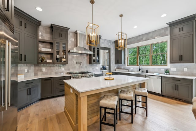 kitchen with a kitchen island, wall chimney range hood, appliances with stainless steel finishes, decorative light fixtures, and light wood-type flooring