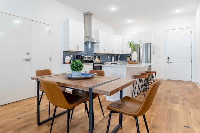 kitchen featuring a center island with sink, white cabinetry, wall chimney range hood, appliances with stainless steel finishes, and light hardwood / wood-style flooring