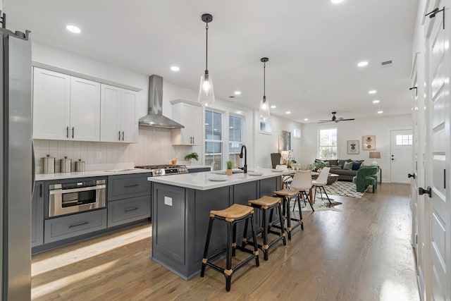 kitchen with white cabinets, hanging light fixtures, an island with sink, wall chimney range hood, and appliances with stainless steel finishes