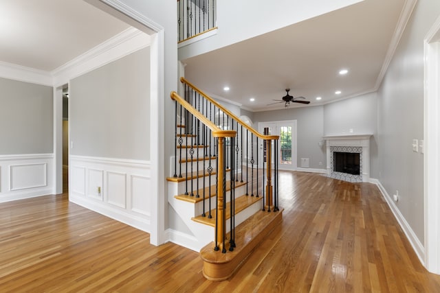 stairway with ceiling fan, wood-type flooring, a tile fireplace, and crown molding