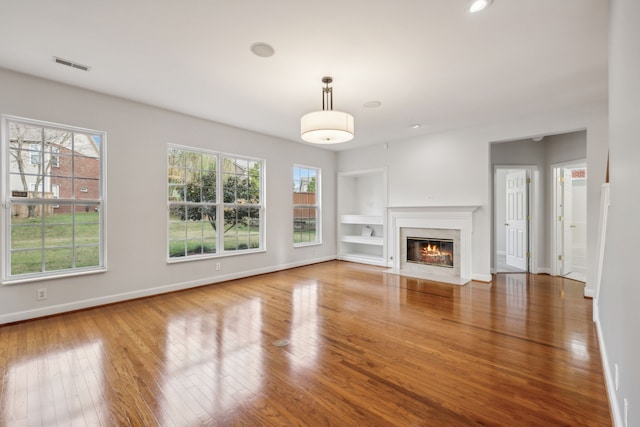 unfurnished living room featuring a fireplace, a wealth of natural light, built in shelves, and hardwood / wood-style flooring