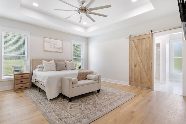 bedroom with ensuite bath, light wood-type flooring, a barn door, a raised ceiling, and ceiling fan