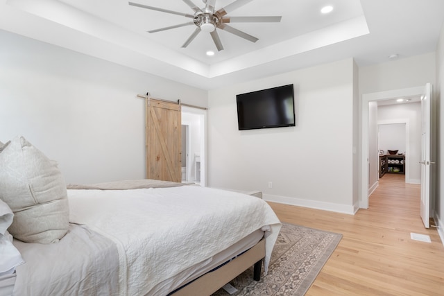 bedroom featuring a barn door, light hardwood / wood-style floors, ceiling fan, and a raised ceiling