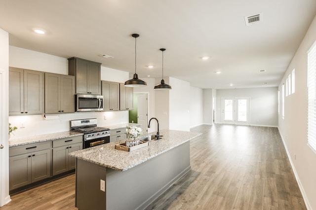 kitchen with stainless steel appliances, light stone countertops, wood-type flooring, a kitchen island with sink, and pendant lighting