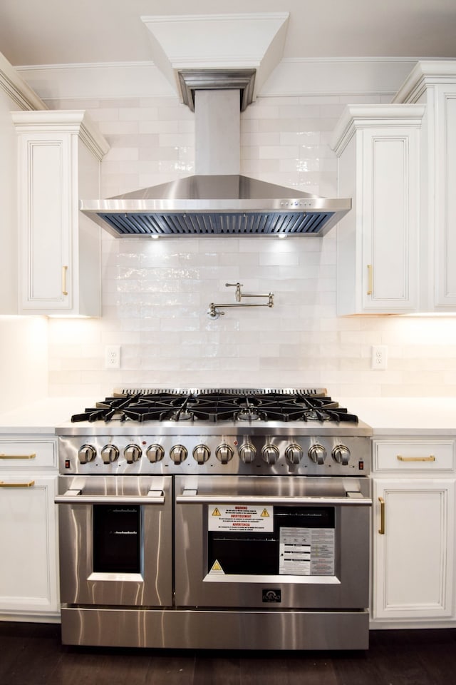 kitchen featuring range with two ovens, light countertops, backsplash, white cabinetry, and wall chimney exhaust hood