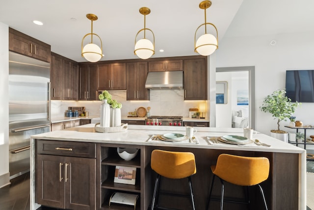 kitchen with extractor fan, appliances with stainless steel finishes, tasteful backsplash, dark brown cabinets, and a kitchen island with sink