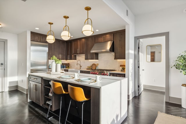 kitchen featuring appliances with stainless steel finishes, backsplash, an island with sink, dark brown cabinets, and pendant lighting