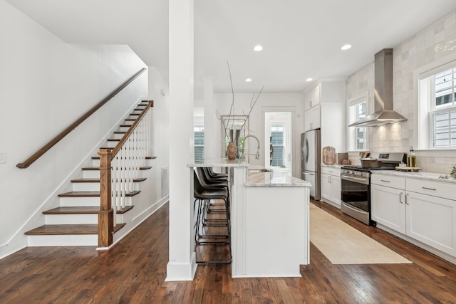 kitchen with stainless steel appliances, white cabinetry, wall chimney range hood, dark hardwood / wood-style floors, and an island with sink