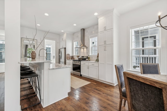 kitchen featuring appliances with stainless steel finishes, a kitchen bar, a kitchen island with sink, white cabinets, and wall chimney range hood