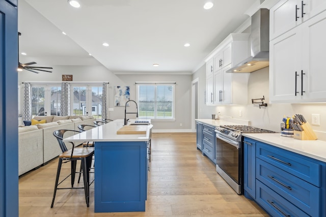 kitchen featuring gas stove, a center island with sink, white cabinetry, wall chimney range hood, and blue cabinetry