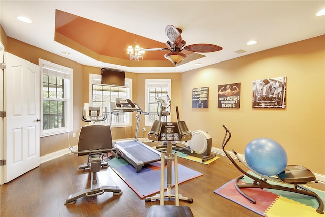 exercise room with dark wood-type flooring, ceiling fan with notable chandelier, and a raised ceiling