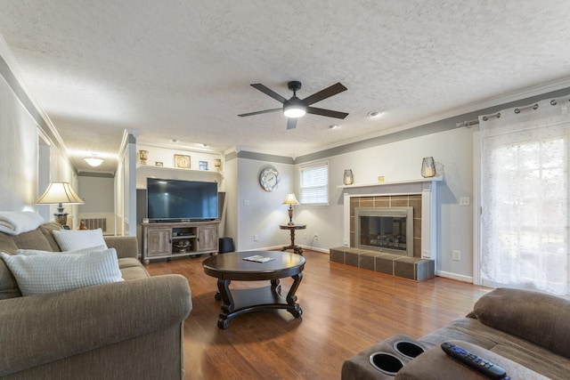 living room featuring hardwood / wood-style floors, a tiled fireplace, ornamental molding, ceiling fan, and a textured ceiling