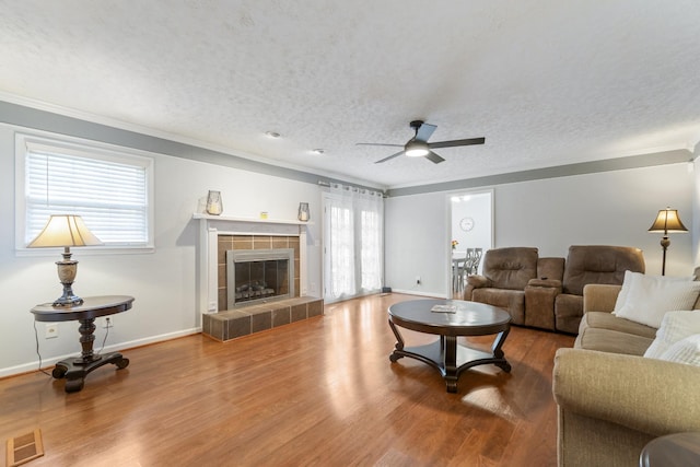 living room featuring crown molding, wood-type flooring, a textured ceiling, a tile fireplace, and ceiling fan