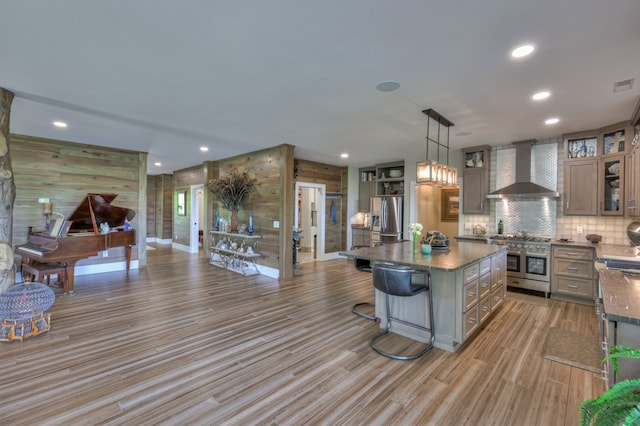 kitchen with stainless steel appliances, wall chimney exhaust hood, wooden walls, a kitchen island, and decorative light fixtures