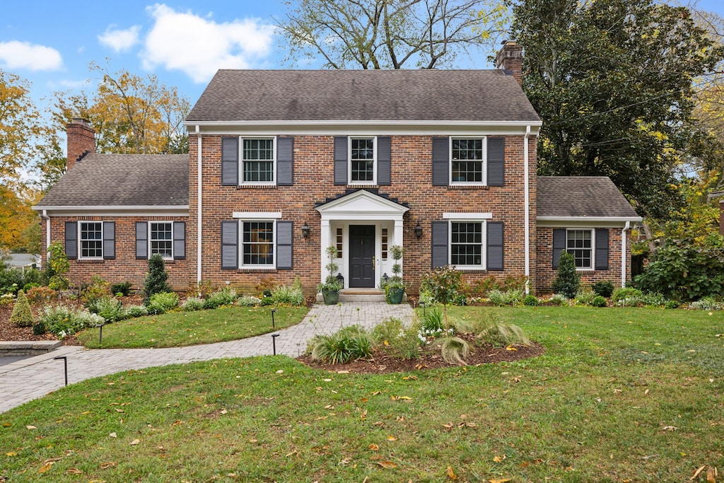 colonial house featuring a shingled roof, brick siding, a chimney, and a front lawn