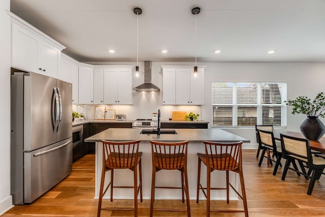 kitchen featuring pendant lighting, wall chimney exhaust hood, light wood-type flooring, an island with sink, and stainless steel refrigerator