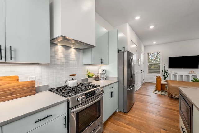 kitchen with decorative backsplash, stainless steel appliances, wall chimney range hood, white cabinets, and light hardwood / wood-style floors