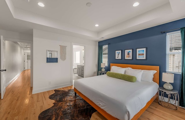 bedroom with light wood-type flooring, connected bathroom, and a raised ceiling