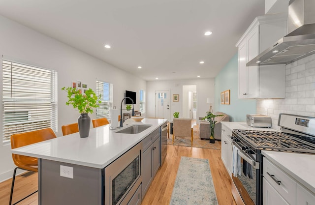 kitchen with a breakfast bar, a center island with sink, white cabinets, wall chimney range hood, and appliances with stainless steel finishes