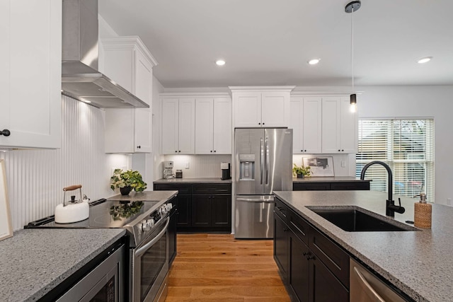 kitchen featuring appliances with stainless steel finishes, light stone counters, wall chimney exhaust hood, sink, and light hardwood / wood-style flooring