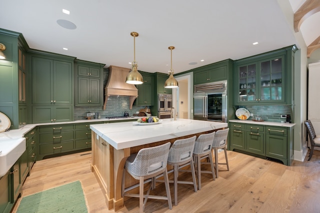 kitchen featuring pendant lighting, light hardwood / wood-style floors, stainless steel built in refrigerator, custom exhaust hood, and a kitchen island with sink