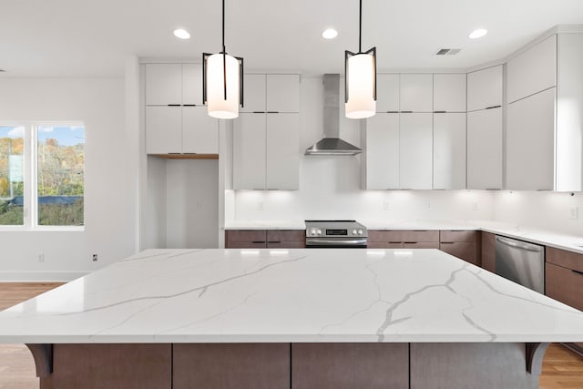 kitchen featuring wall chimney range hood, appliances with stainless steel finishes, decorative light fixtures, a kitchen island, and light wood-type flooring