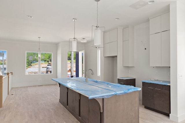 kitchen featuring light stone counters, decorative light fixtures, an island with sink, white cabinets, and light hardwood / wood-style flooring