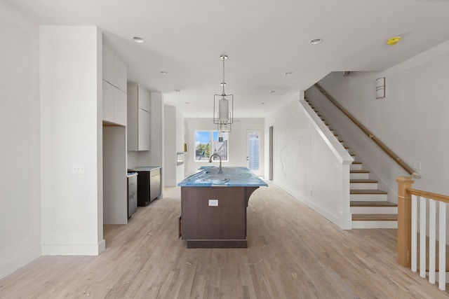 kitchen with a center island with sink, hanging light fixtures, sink, white cabinetry, and light wood-type flooring