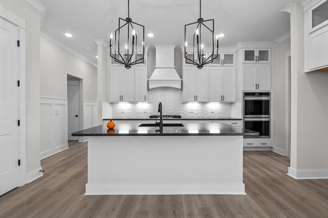 kitchen featuring a center island with sink, white cabinets, double oven, decorative light fixtures, and premium range hood