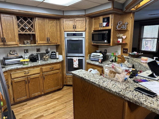 kitchen with stainless steel appliances, a paneled ceiling, light stone countertops, backsplash, and light hardwood / wood-style flooring