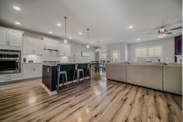 kitchen featuring white cabinets, a kitchen breakfast bar, hanging light fixtures, an island with sink, and double oven