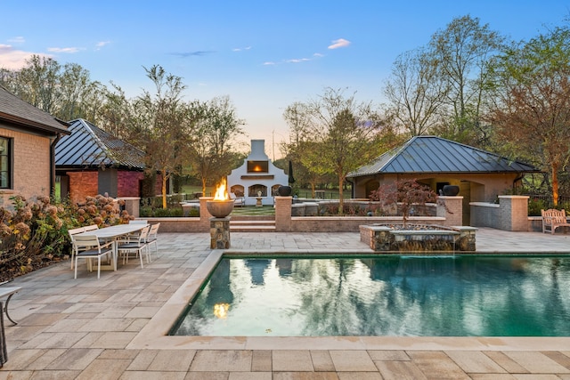 pool at dusk with an outdoor fireplace, pool water feature, and a patio