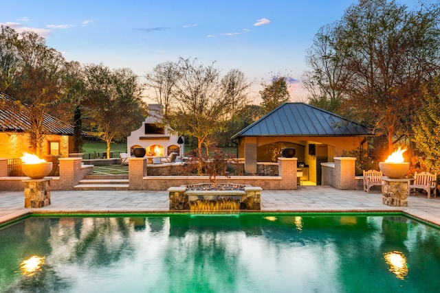 pool at dusk with exterior fireplace, a patio, and pool water feature