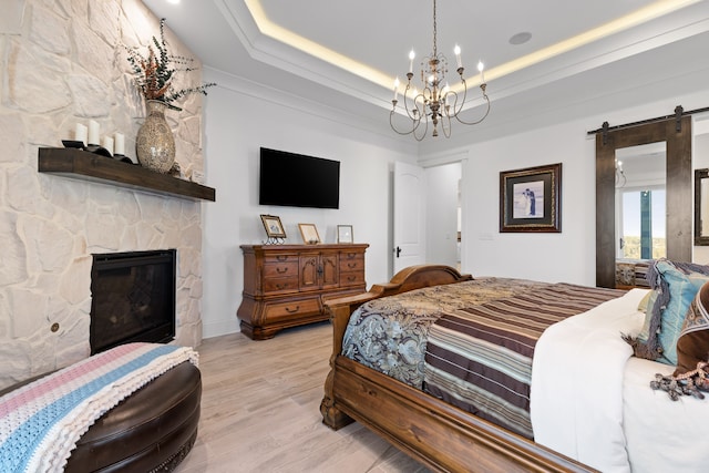 bedroom with a stone fireplace, a notable chandelier, a barn door, a tray ceiling, and light wood-type flooring