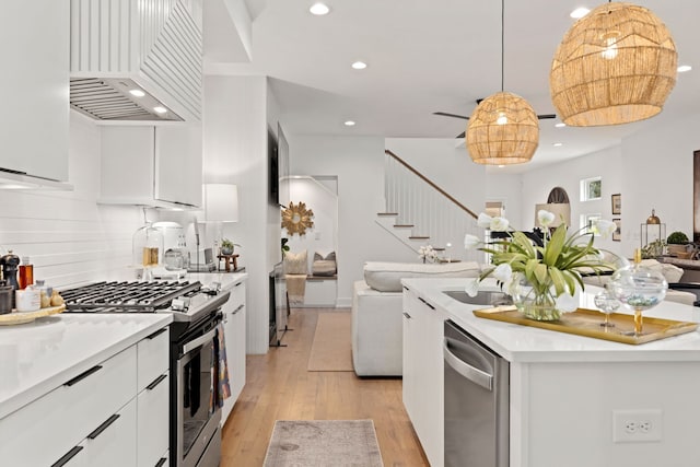 kitchen featuring appliances with stainless steel finishes, custom range hood, hanging light fixtures, white cabinets, and light hardwood / wood-style flooring