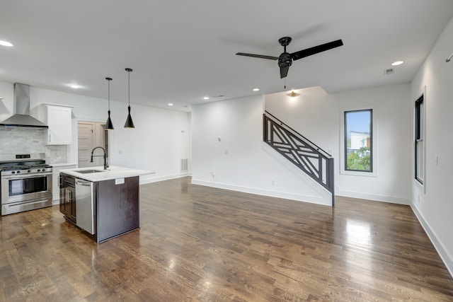 kitchen featuring stainless steel appliances, a center island with sink, white cabinetry, decorative light fixtures, and wall chimney range hood