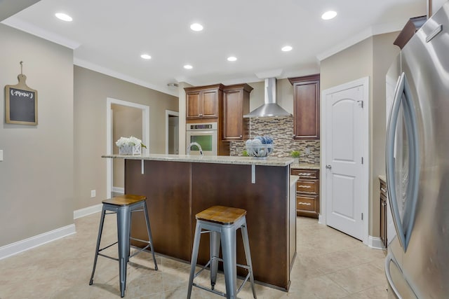 kitchen featuring appliances with stainless steel finishes, light stone countertops, a kitchen bar, an island with sink, and wall chimney range hood