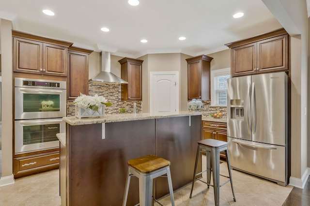 kitchen with wall chimney range hood, appliances with stainless steel finishes, light stone countertops, a breakfast bar area, and a center island