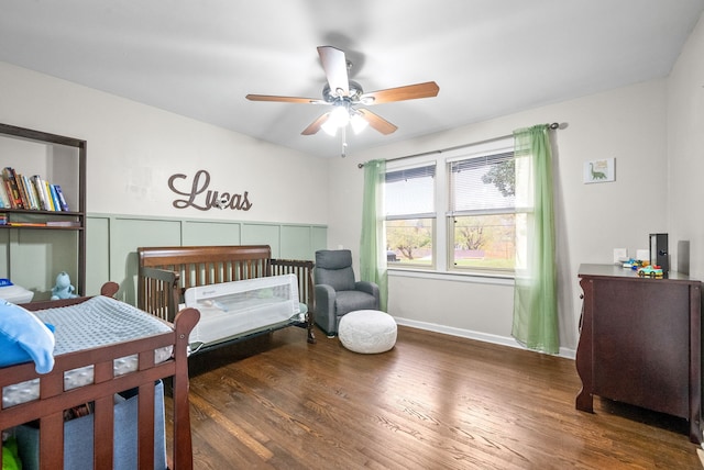 bedroom with dark wood-type flooring, a nursery area, and ceiling fan