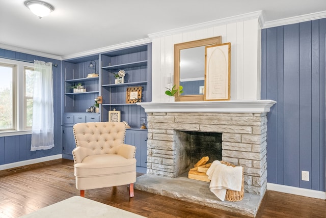 sitting room with ornamental molding, built in shelves, dark wood-type flooring, and a stone fireplace