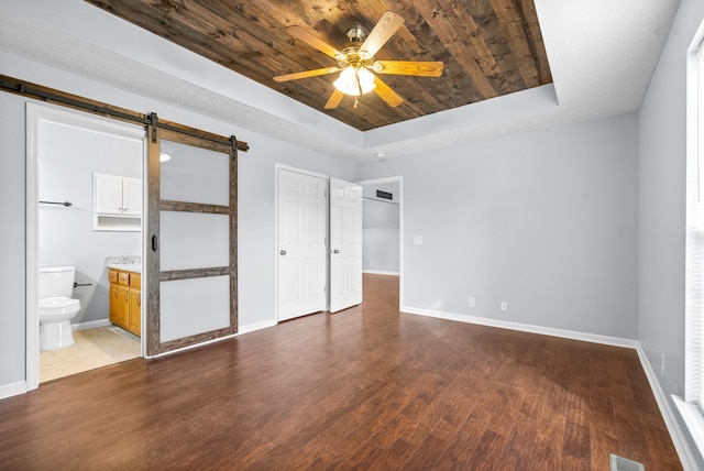 unfurnished bedroom featuring ceiling fan, hardwood / wood-style floors, a raised ceiling, a barn door, and connected bathroom