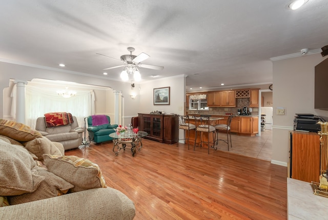 living room featuring light wood-type flooring, ceiling fan with notable chandelier, crown molding, and decorative columns