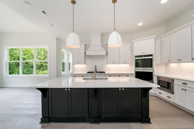 kitchen featuring white cabinetry, hanging light fixtures, premium range hood, an island with sink, and appliances with stainless steel finishes