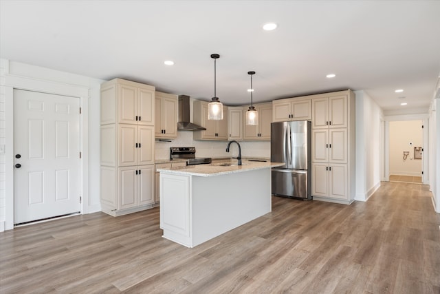 kitchen featuring wall chimney range hood, light hardwood / wood-style flooring, an island with sink, pendant lighting, and appliances with stainless steel finishes