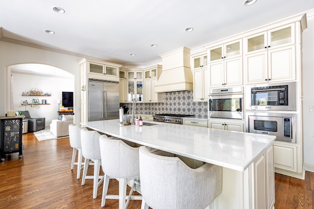 kitchen featuring dark wood-style flooring, custom range hood, backsplash, a sink, and built in appliances