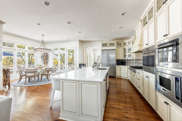 kitchen with a center island with sink, arched walkways, dark wood-style floors, built in appliances, and backsplash