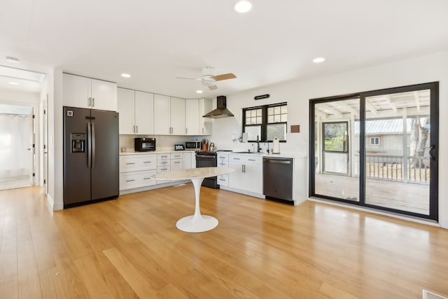 kitchen featuring light wood-type flooring, wall chimney range hood, sink, black appliances, and white cabinetry