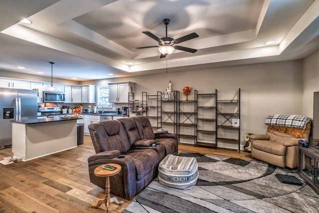 living room with a raised ceiling, ceiling fan, sink, and light wood-type flooring