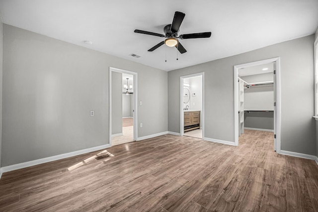 unfurnished bedroom with ensuite bath, a spacious closet, a closet, ceiling fan with notable chandelier, and light wood-type flooring