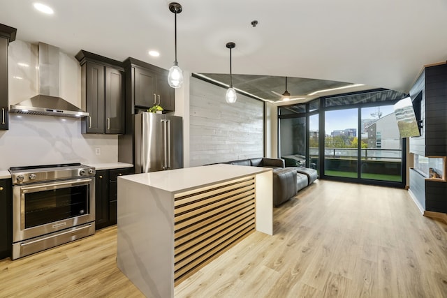 kitchen featuring wall chimney range hood, light wood-type flooring, decorative light fixtures, a kitchen island, and stainless steel appliances